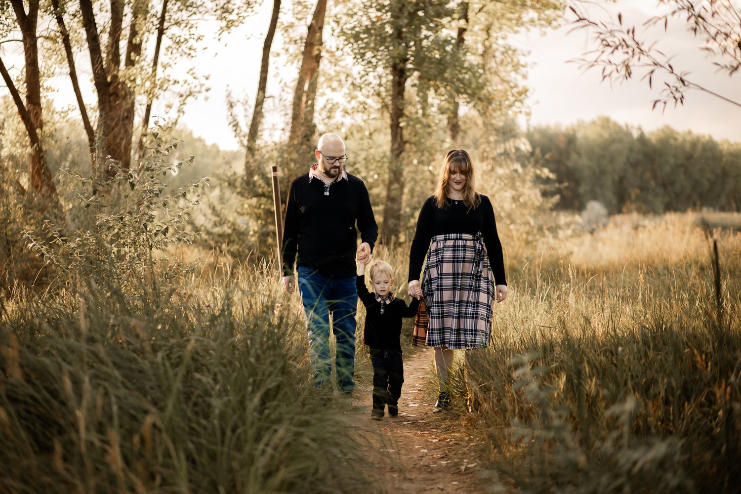 family of 3 wearing Fall colors posing for a picture walking down a dirt road in a forest setting. Liz Rivas with Illuminating Images photog