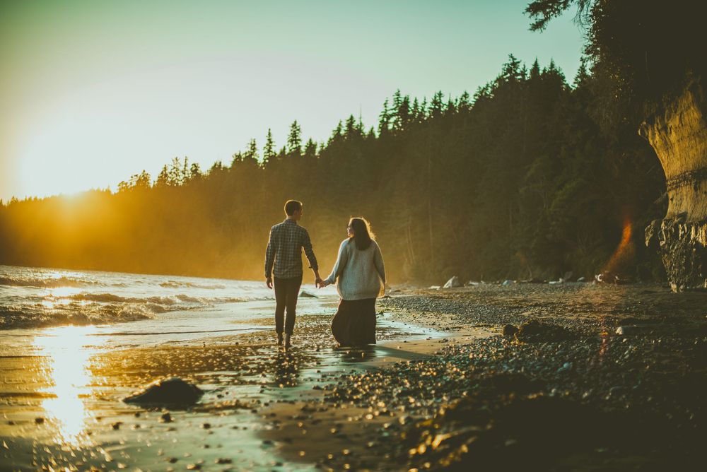 Couple hold hands while walking along a beach at sunset with light casting golden reflections on the water Edmonton Engagement Photographer