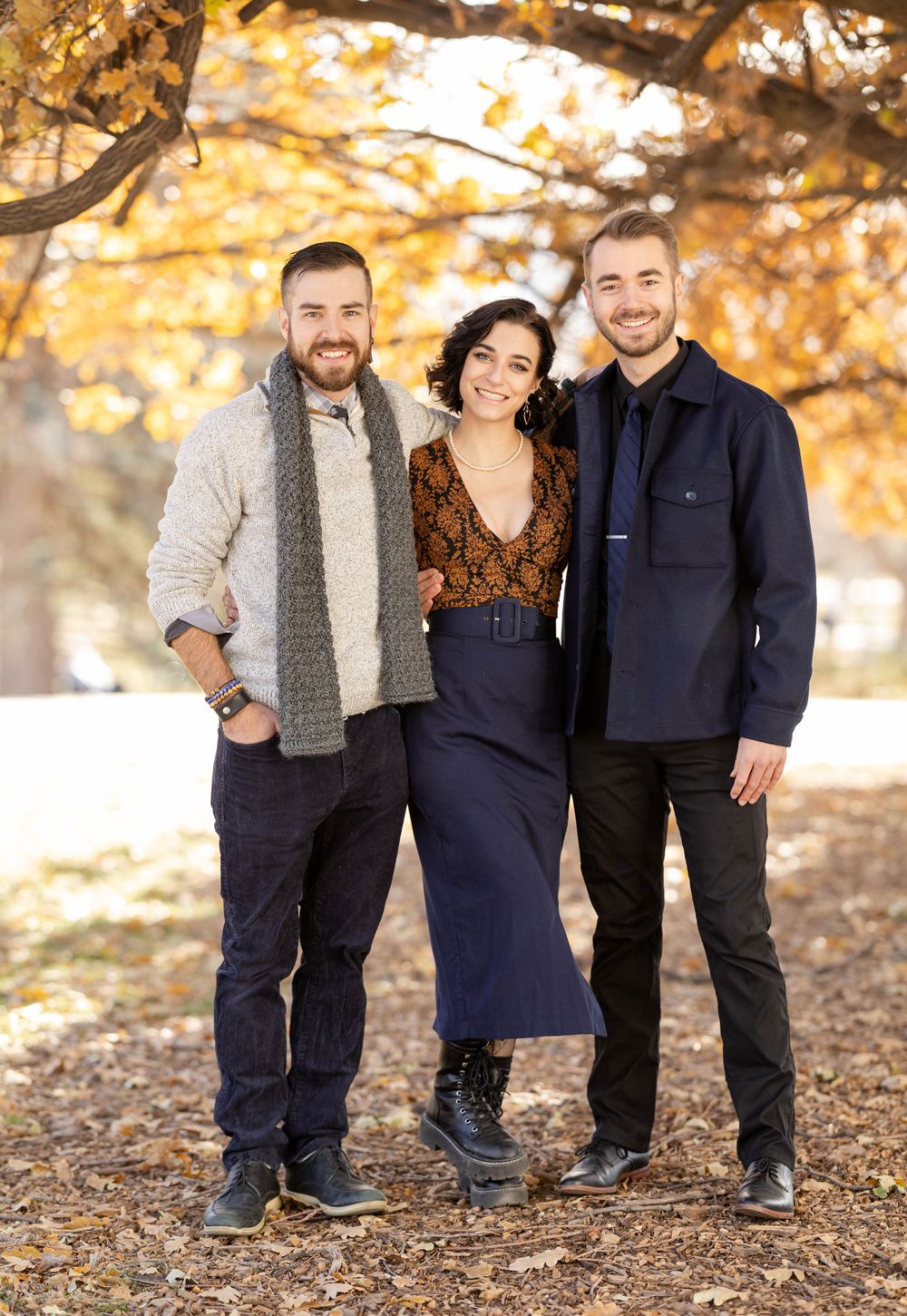 Three people posing together under autumn trees wearing casual stylish outfits.