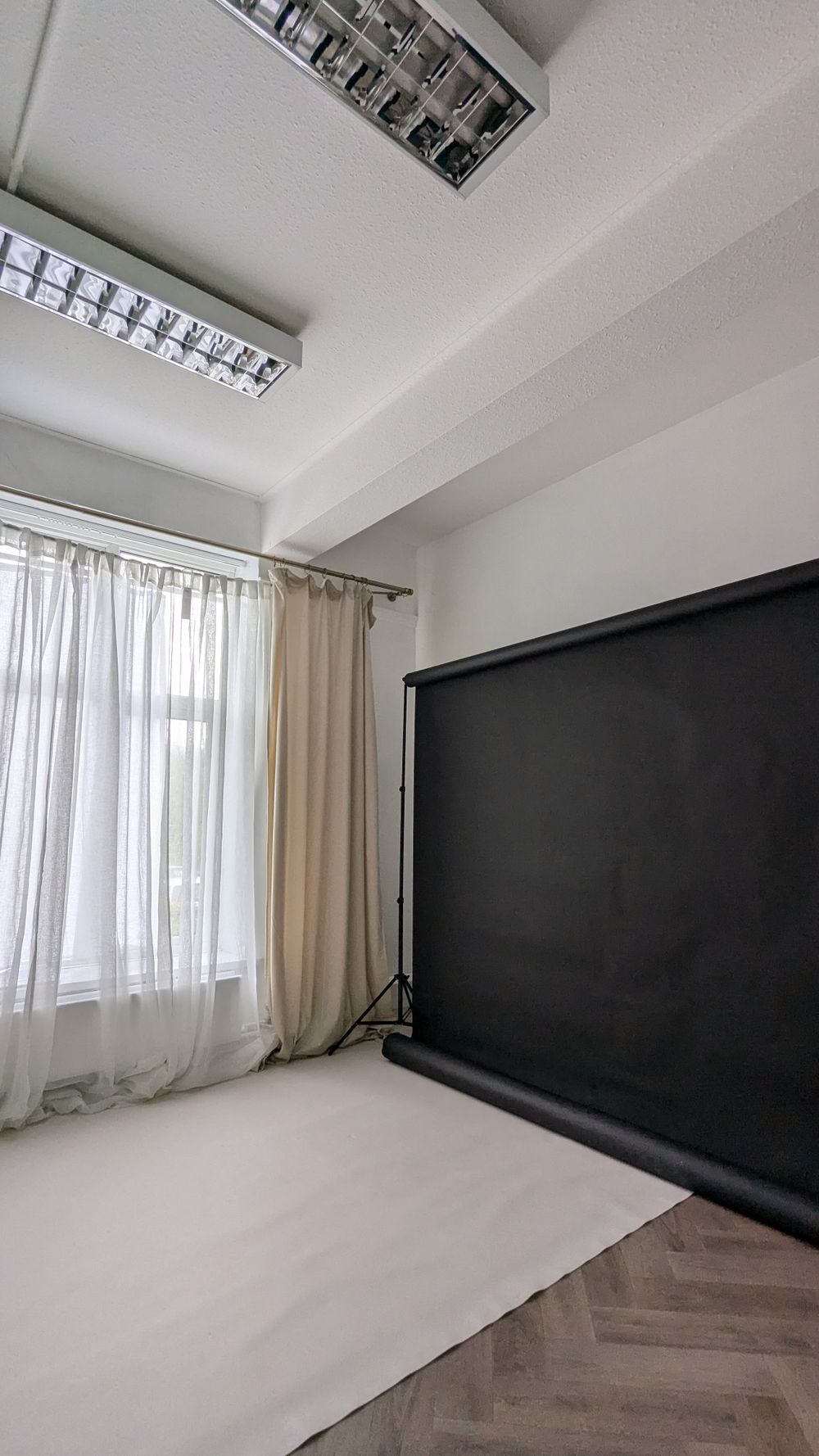 Interior studio room with black backdrop, white curtains, wooden floor and fluorescent ceiling lights.