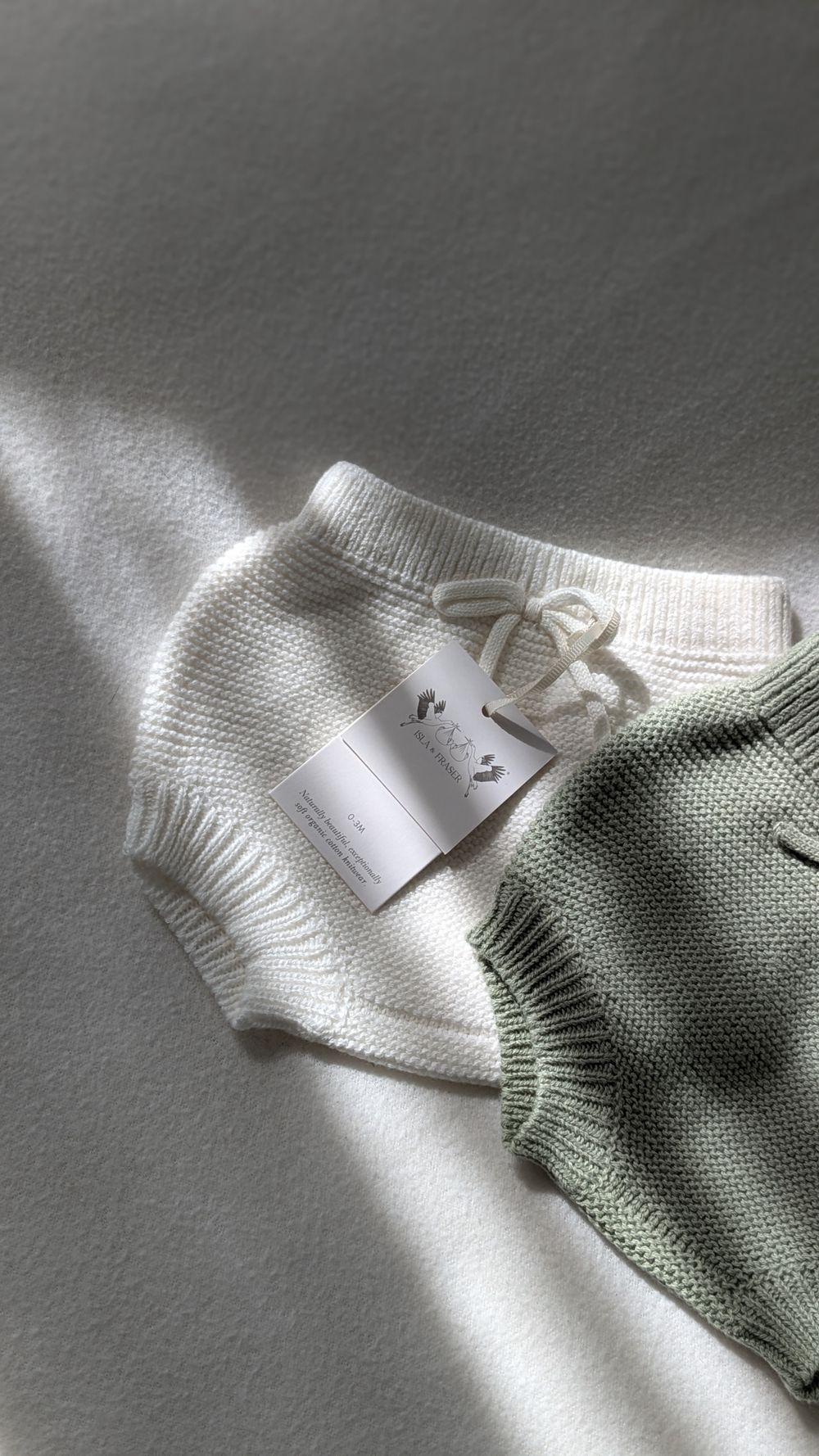Soft knitted sweaters in white and sage green with clothing tag, captured in gentle natural lighting.