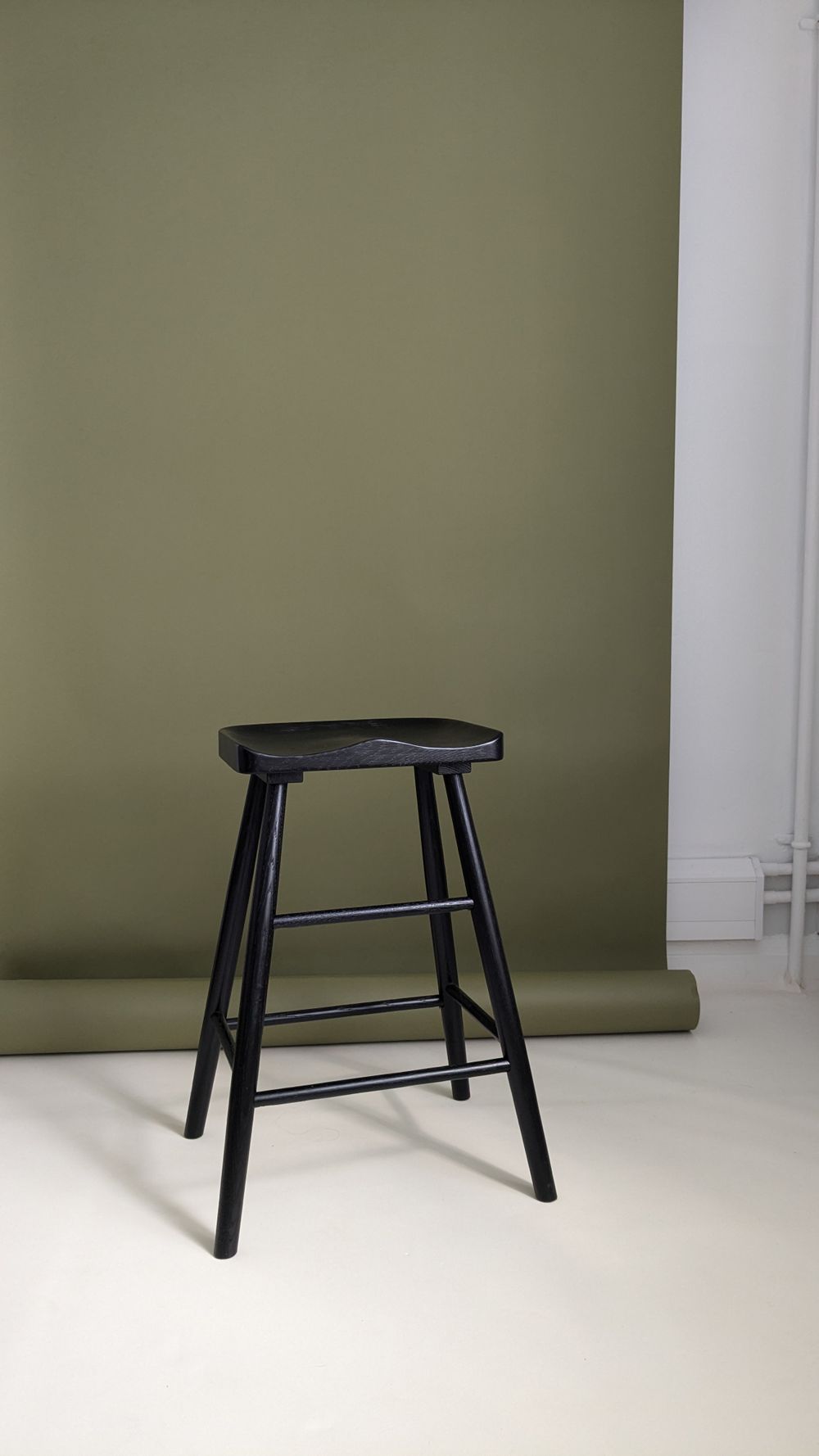 A minimalist black wooden stool stands against an olive green wall backdrop with white flooring.