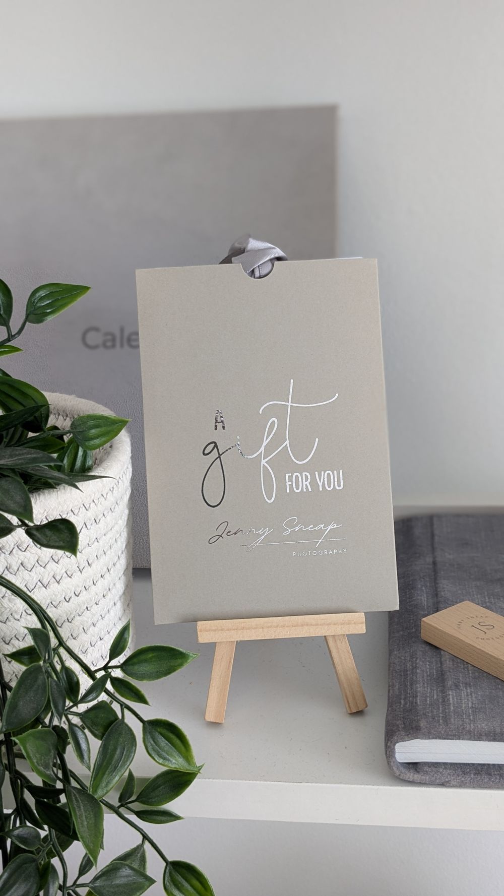 A small wooden easel displays a beige greeting card with "Gift for you" text next to a potted plant and notebook.