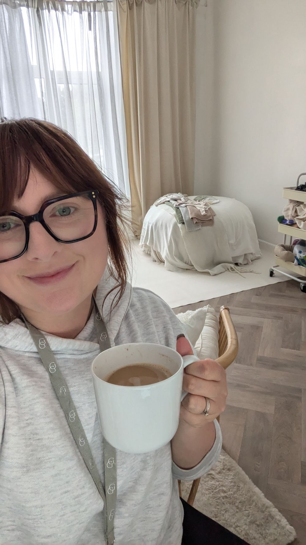 A cozy morning selfie showing a warm cup of coffee in a white mug with curtained windows and home decor in the background.