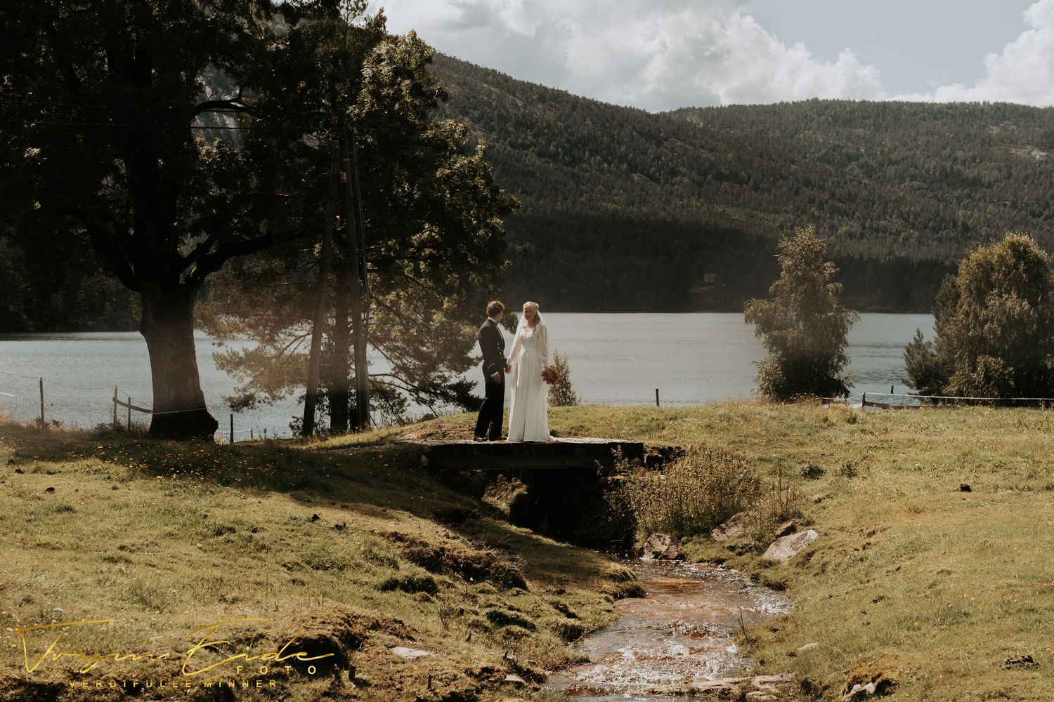 A couple stands on a hillside overlooking a serene lake during an outdoor wedding ceremony on a cloudy day.