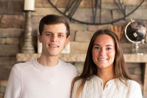 Two young people stand close together in front of a stone wall with a large clock display.