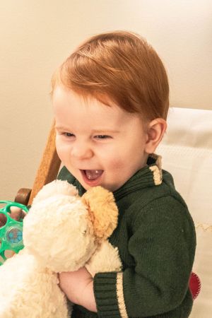 Laughing while hugging a cream colored plush toy in a green knit sweater.
