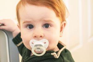 A toddler with bright eyes and a pacifier wearing a dark green sweater.