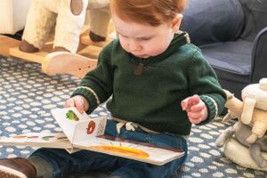 A small child in a green sweater explores a book while sitting on a patterned floor.