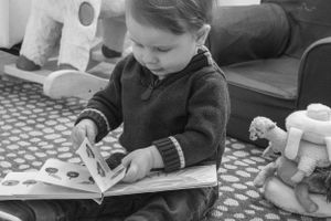 A small child concentrating on reading a book while sitting on a patterned rug.