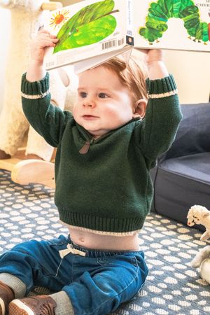 A small child in a green sweater holds up a book featuring colorful illustrations.
