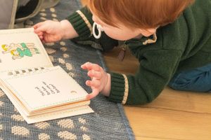 A small reader in green studies pages of a book while lying on a patterned rug.