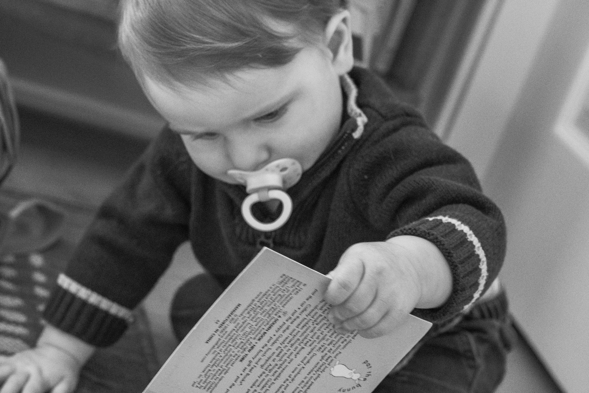 A toddler with a pacifier reads a book while wearing a dark sweater.