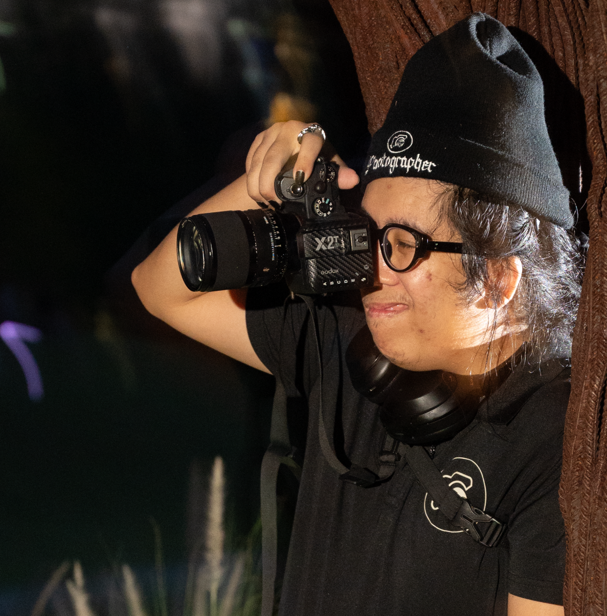 Photographer wearing a black beanie and glasses holding a professional camera in low light.