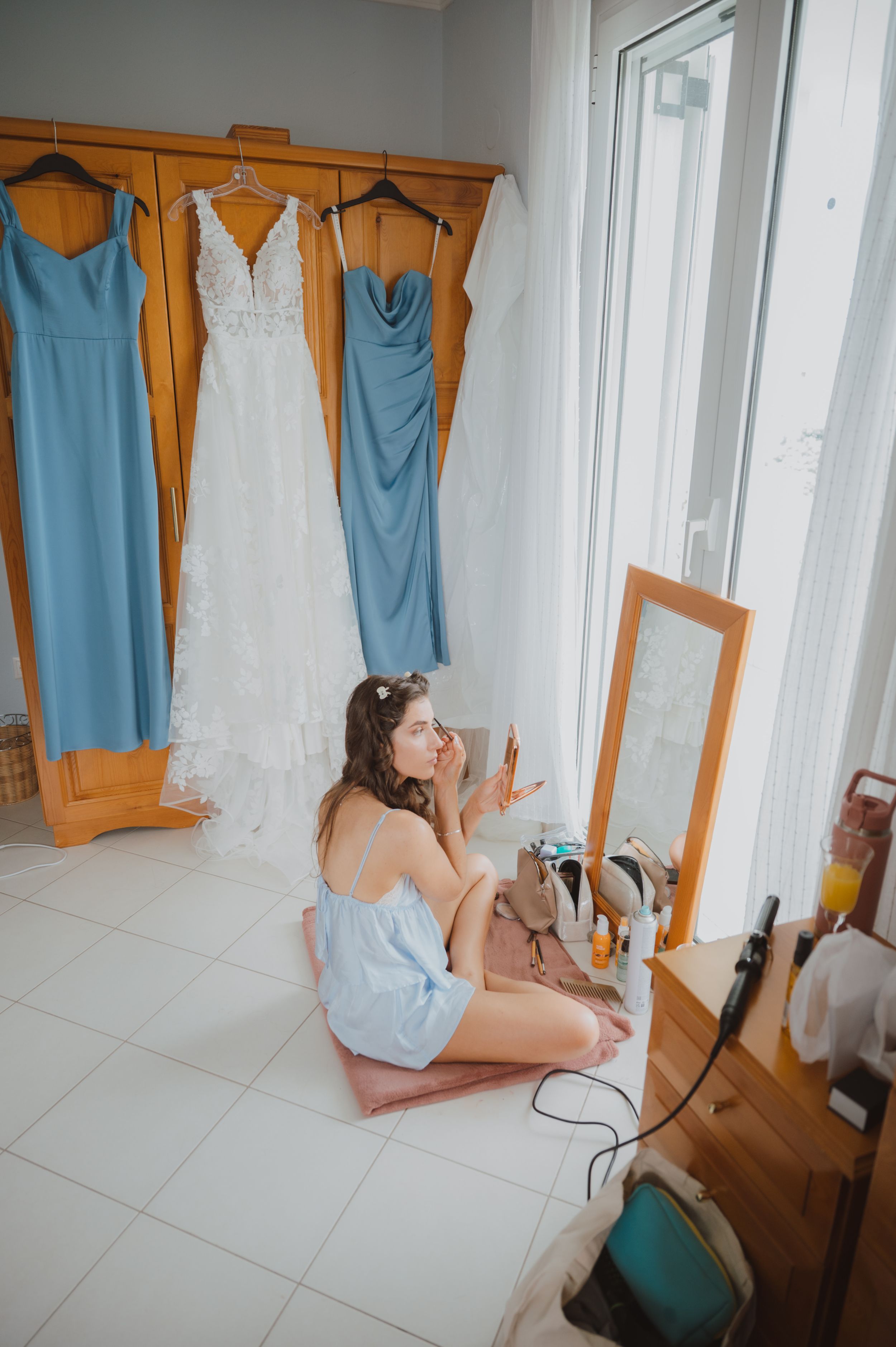 Light blue bridesmaid dresses and a white wedding gown hang on wooden hangers in a bright room.