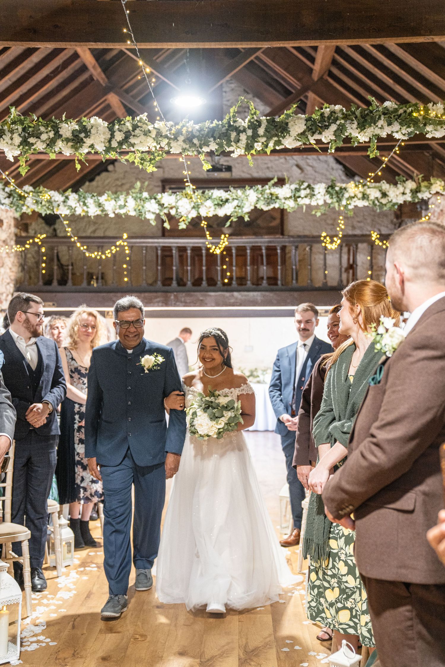 A wedding ceremony in a rustic barn venue decorated with white floral garlands and twinkling lights overhead.