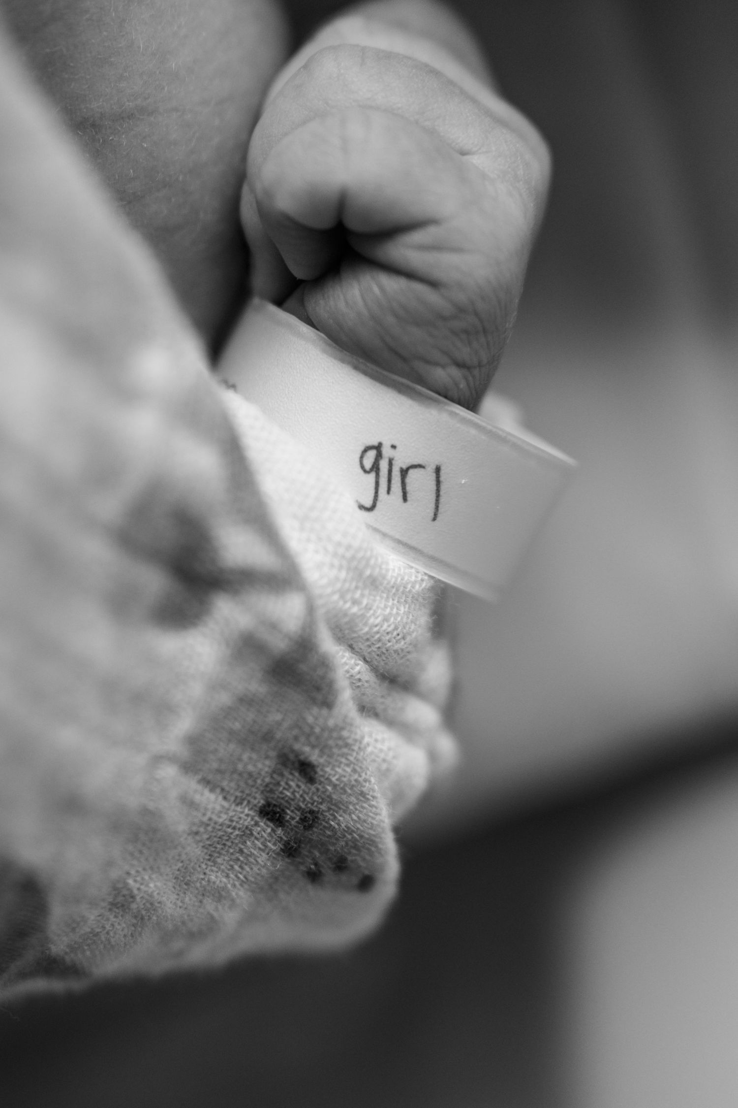 Black and white photo of a newborn baby girl's tiny fist with a hospital identification bracelet labeled girl. UPMC Magee Fresh48 Session