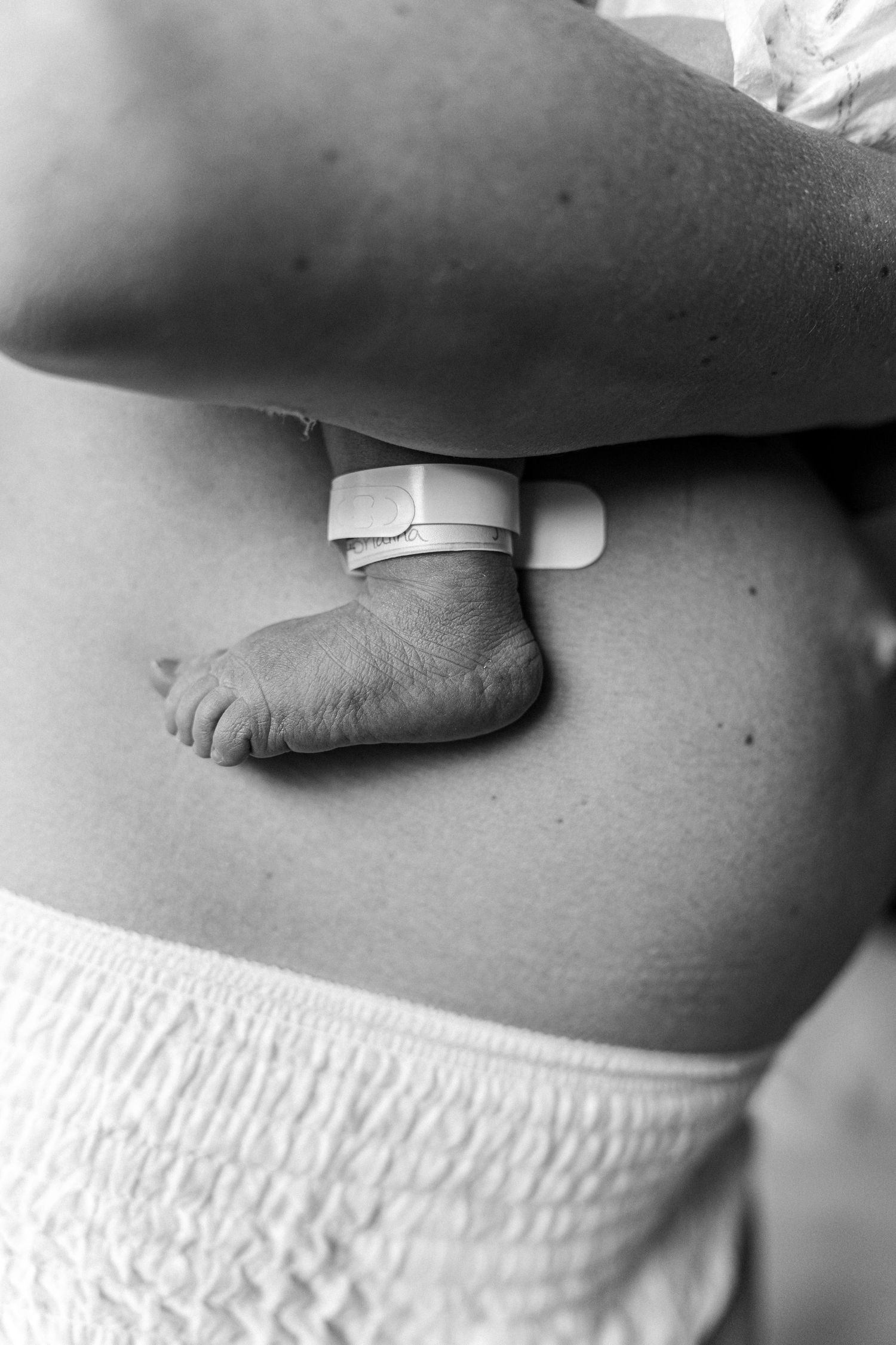 Black and white photo of a newborn baby foot with hospital ID bracelet resting against mother's body. UPMC Magee Fresh48 Session