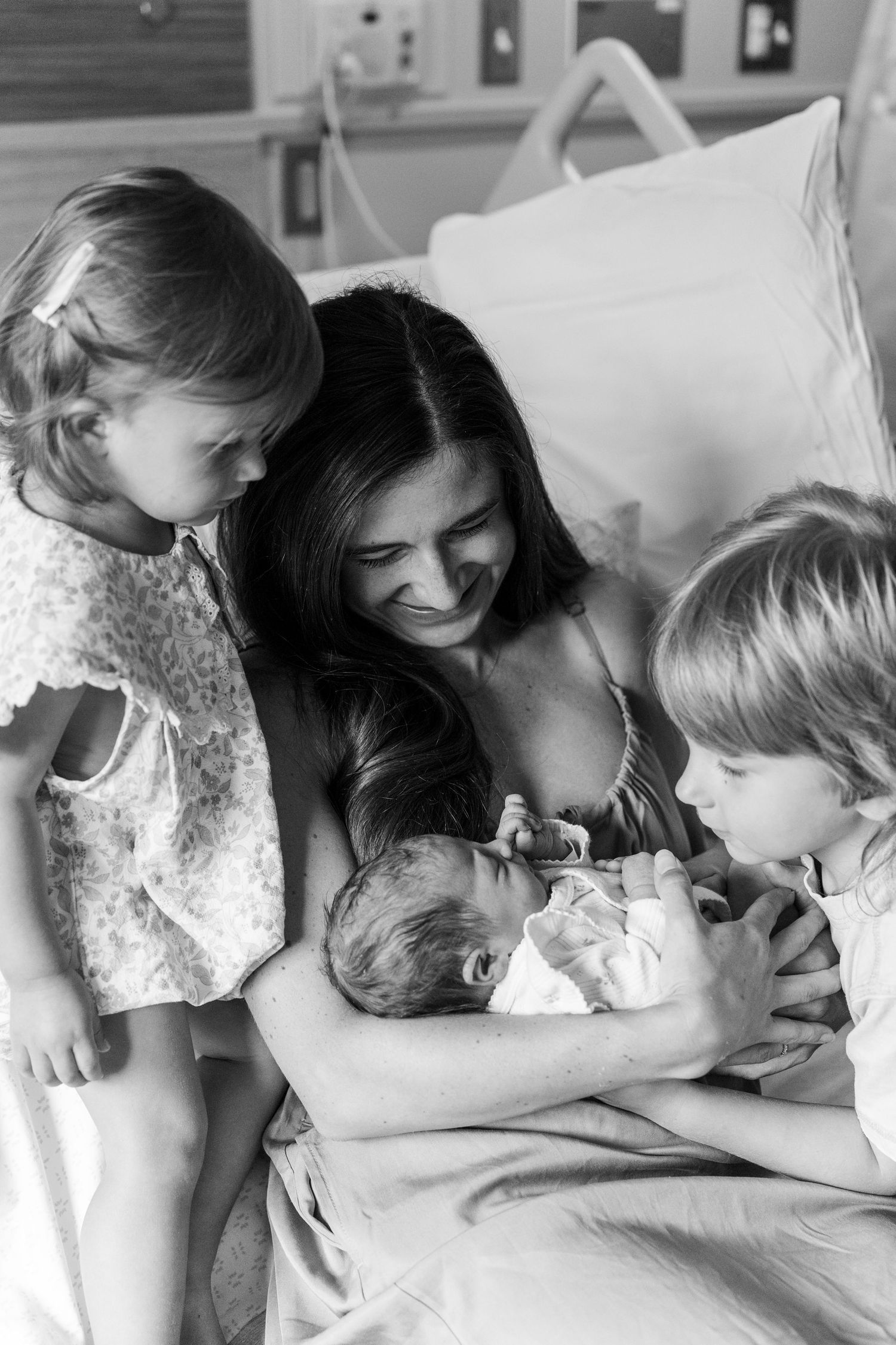 Black and white photo of a mother in hospital bed smiling at newborn baby surrounded by two young siblings. UPMC Magee Fresh48 Session
