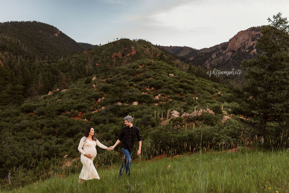 Couple holding hands and walking through scenic mountain meadow with evergreen trees and red rock formations in background.