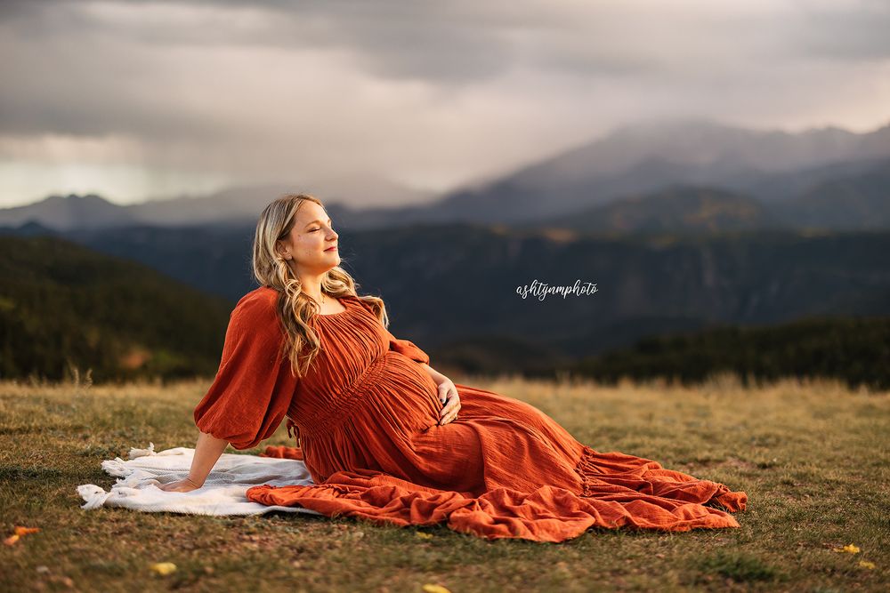 A person in a flowing red dress reclines on a blanket in a mountain meadow at sunset.