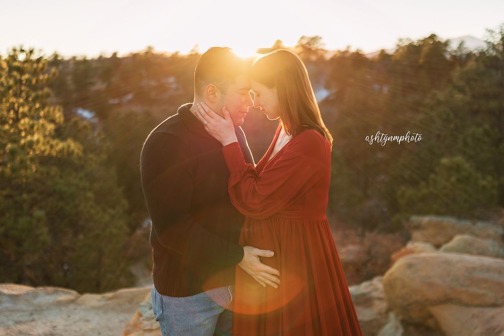 A romantic couple embraces on a rocky overlook at sunset with golden sunlight streaming through the trees.