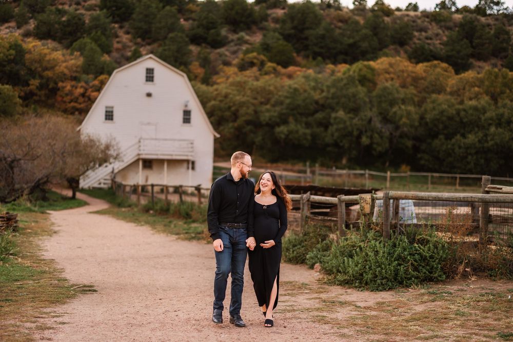 A romantic couple walks together on a dirt path near a rustic white barn during golden hour.