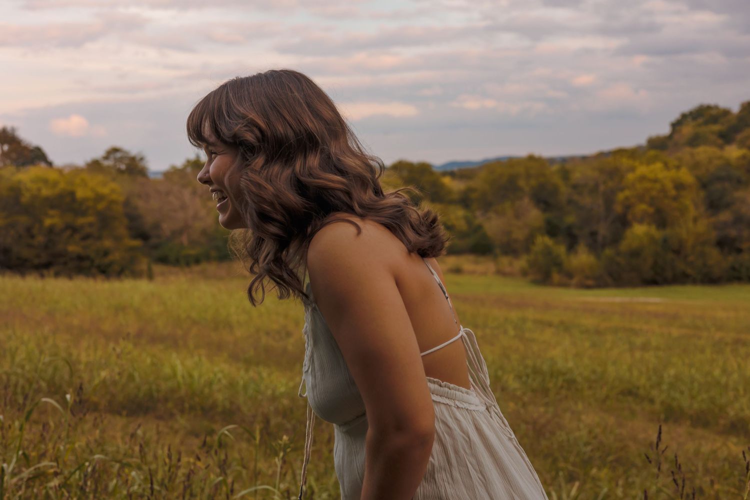 A woman in a white dress stands in a golden field at sunset with rolling hills in the background.