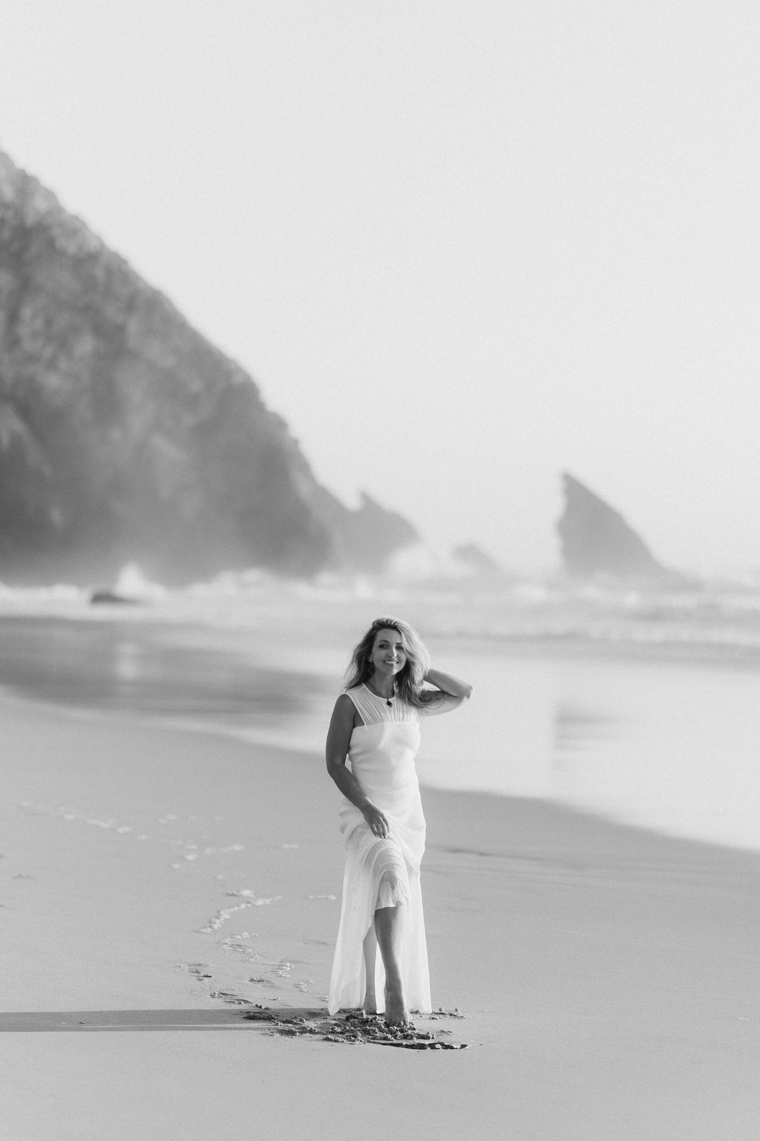 Woman in white dress standing on a misty beach with rocky cliffs, black and white photography.