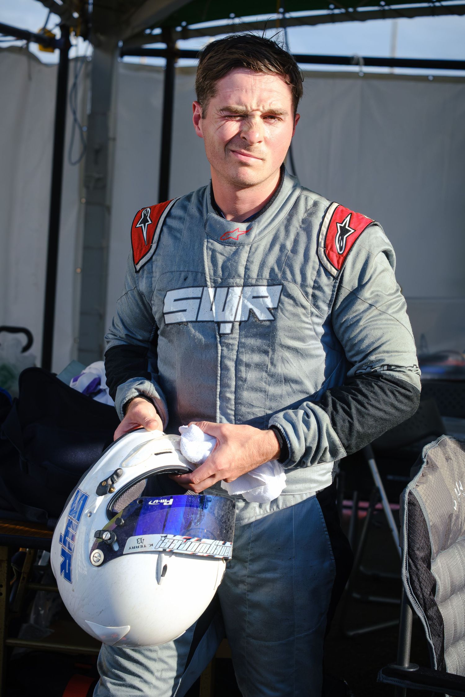 A race car driver in a gray STR racing suit holds a white helmet with blue accents at a motorsport event.