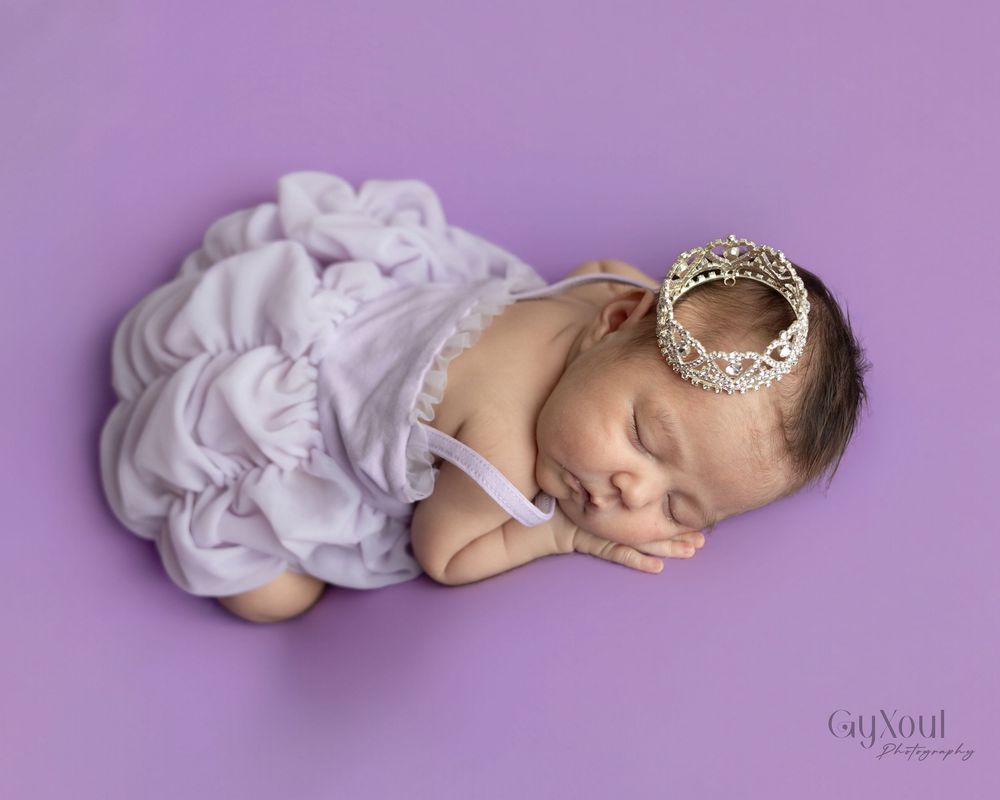 A newborn baby in a white dress sleeps peacefully on a purple backdrop during a professional photography session.