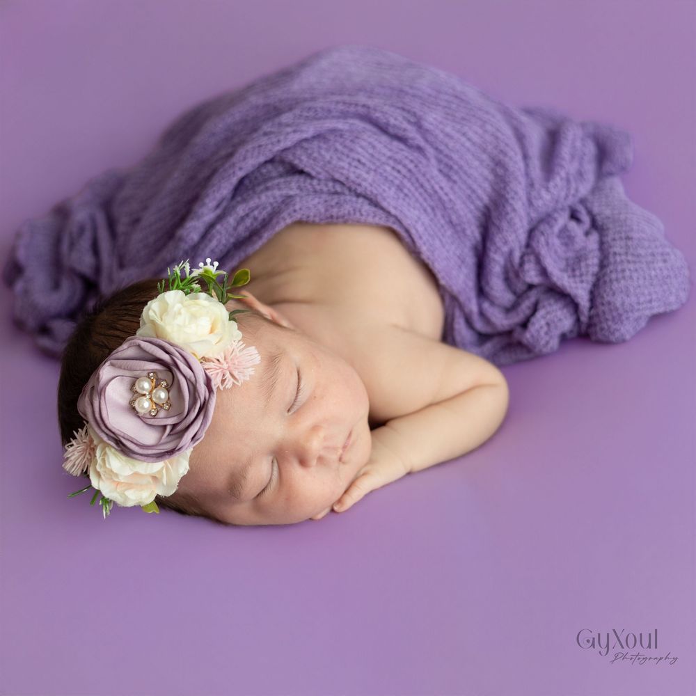 Sleeping newborn wrapped in purple fabric with floral headband on purple background.