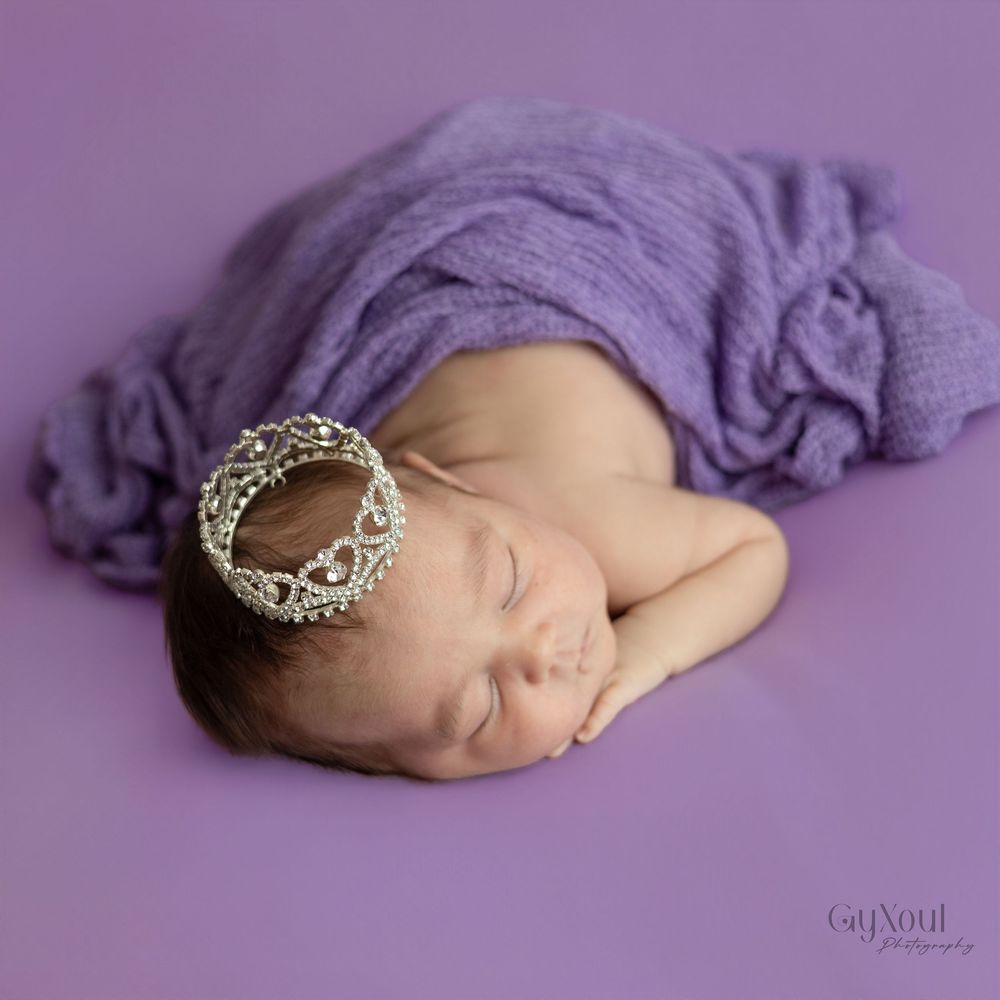 Sleeping newborn wrapped in soft purple fabric with delicate lace headband on purple background.