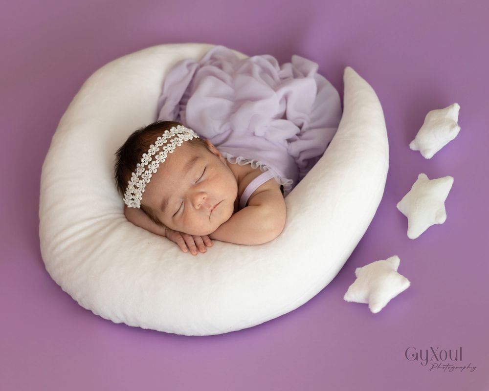 A sleeping newborn baby wearing purple clothing rests peacefully on a white circular pillow with soft toys nearby.