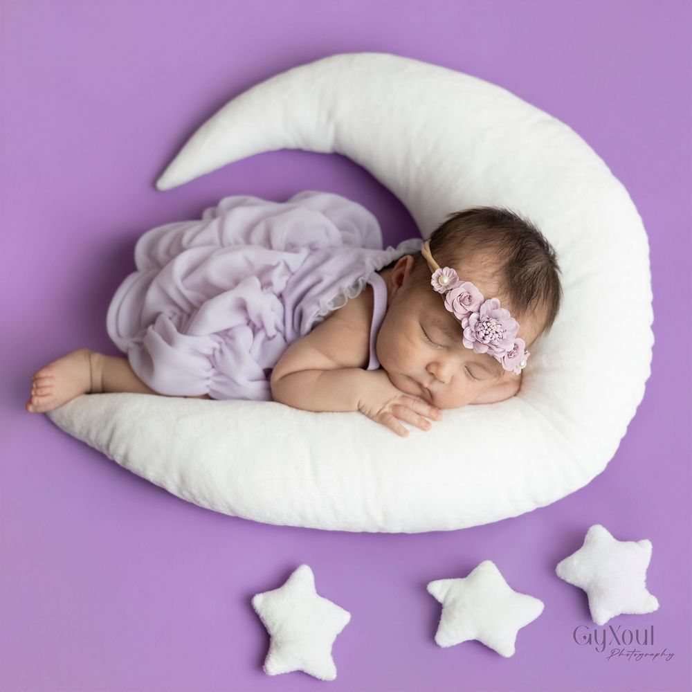 Sleeping baby on white crescent-shaped nursing pillow with star cushions against purple background.