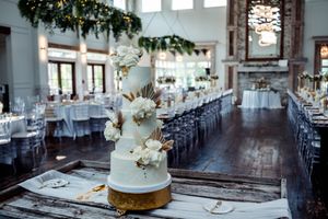 An elegant white wedding cake adorned with flowers sits on a rustic table in a reception hall with long dining tables.