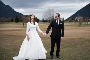 Couple in wedding attire holds hands while walking through a scenic mountain meadow on an overcast day.