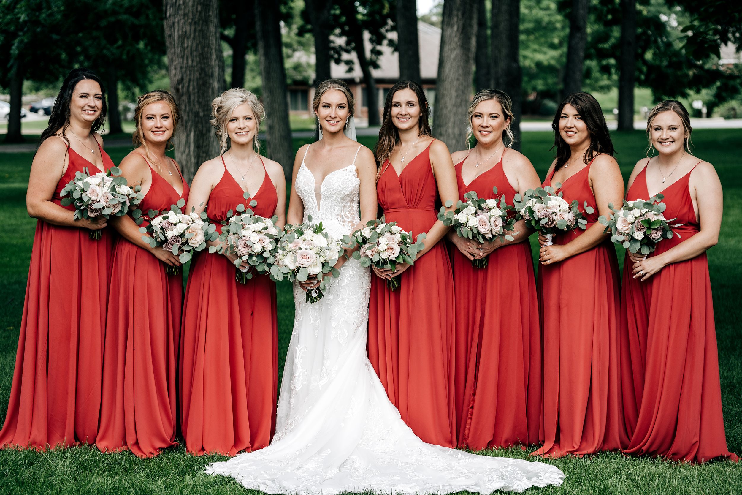 Bridal party in matching floor-length coral red dresses poses with bride in white gown holding floral bouquets outdoors.