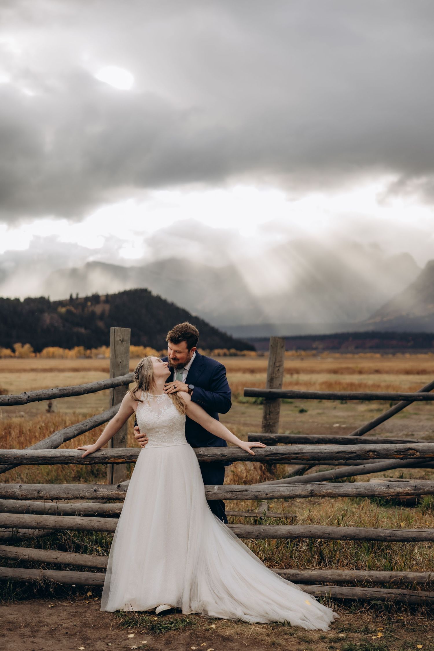 A romantic wedding photo session by a rustic wooden fence in a field with dramatic cloudy skies.