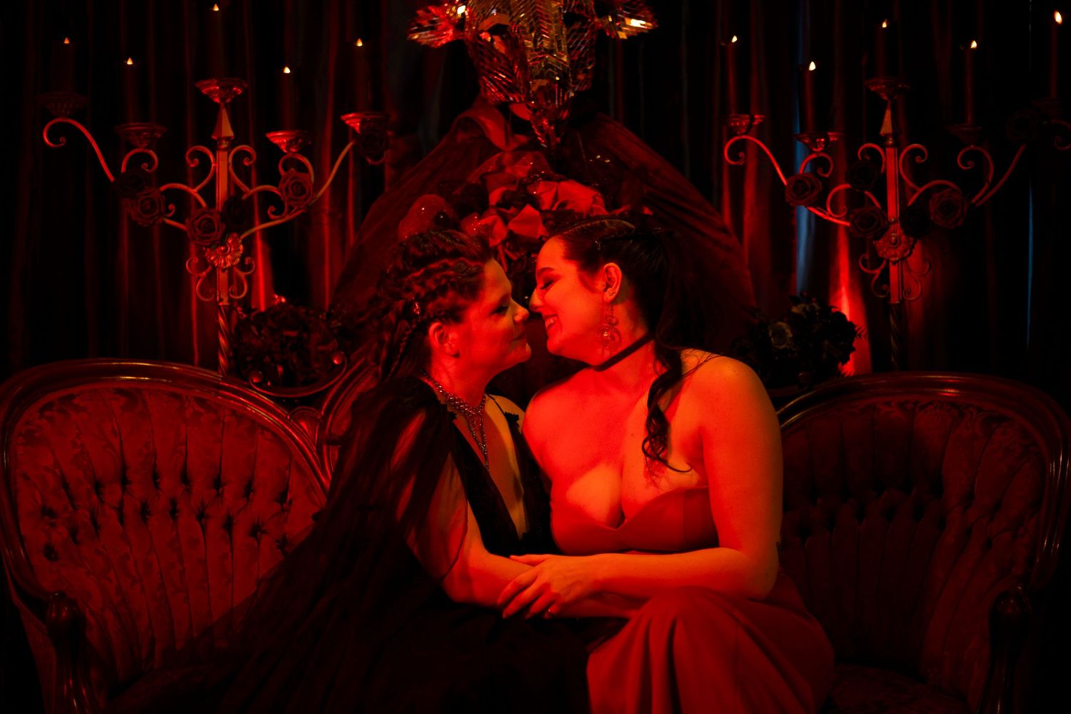 A romantic couple embraces in a dimly lit room bathed in dramatic red lighting with ornate decor in the background.