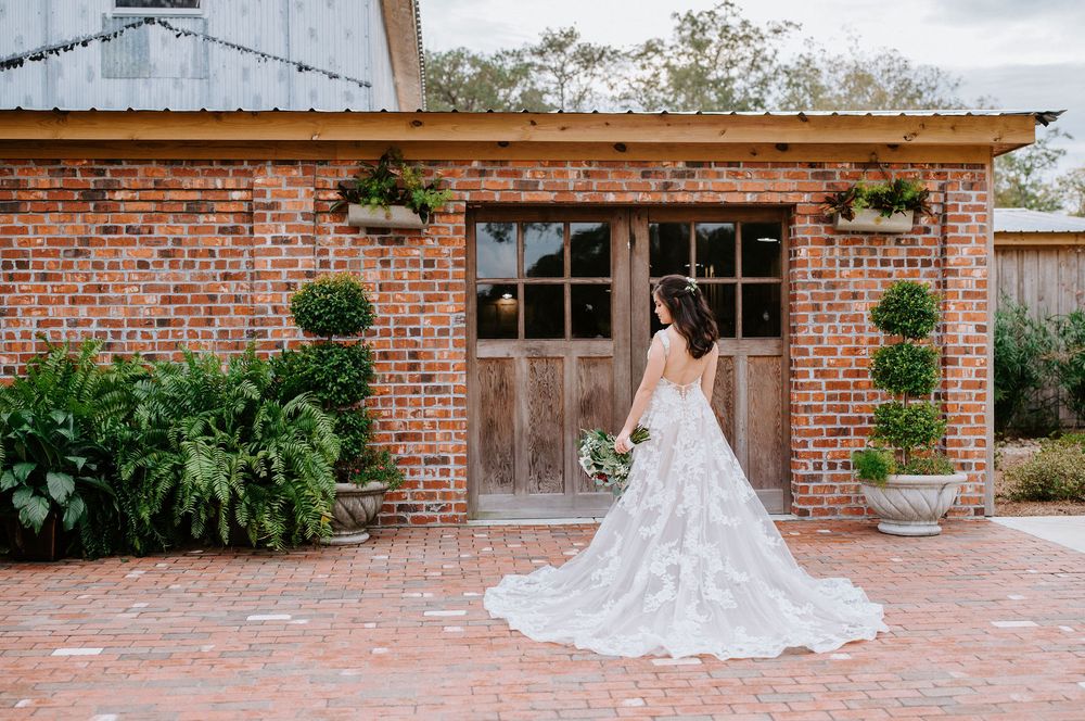 A bride in a flowing lace wedding dress stands before rustic wooden doors at a brick venue with potted topiaries.