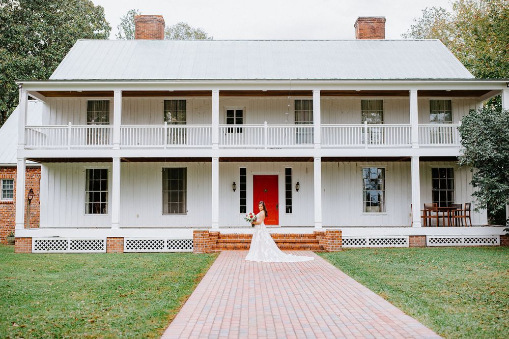 A long white farmhouse with a wraparound porch and brick walkway sits nestled among green trees.