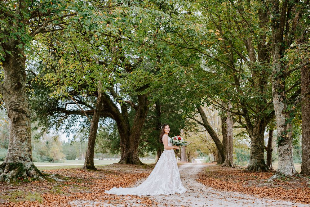 A white wedding dress flows elegantly along a tree-lined autumn path covered in fallen leaves.
