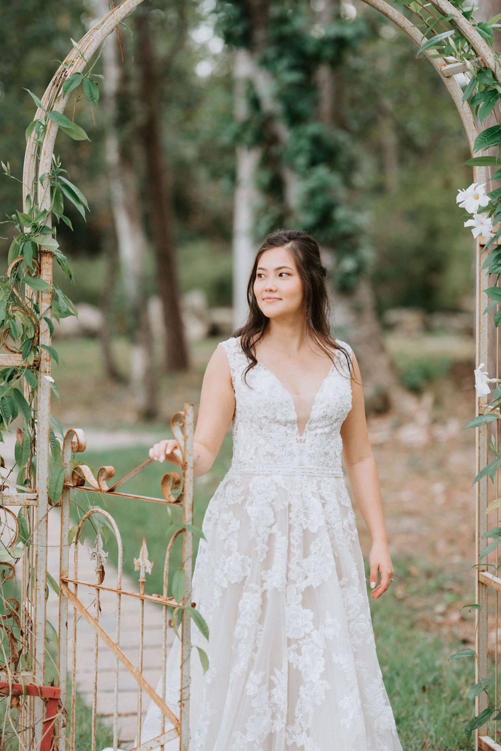 A bride in a white lace dress poses in a garden setting with decorative arches and greenery.