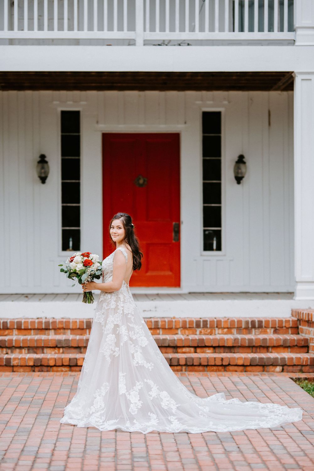 A stunning wedding dress is showcased against a white house with a bold red door and brick steps.