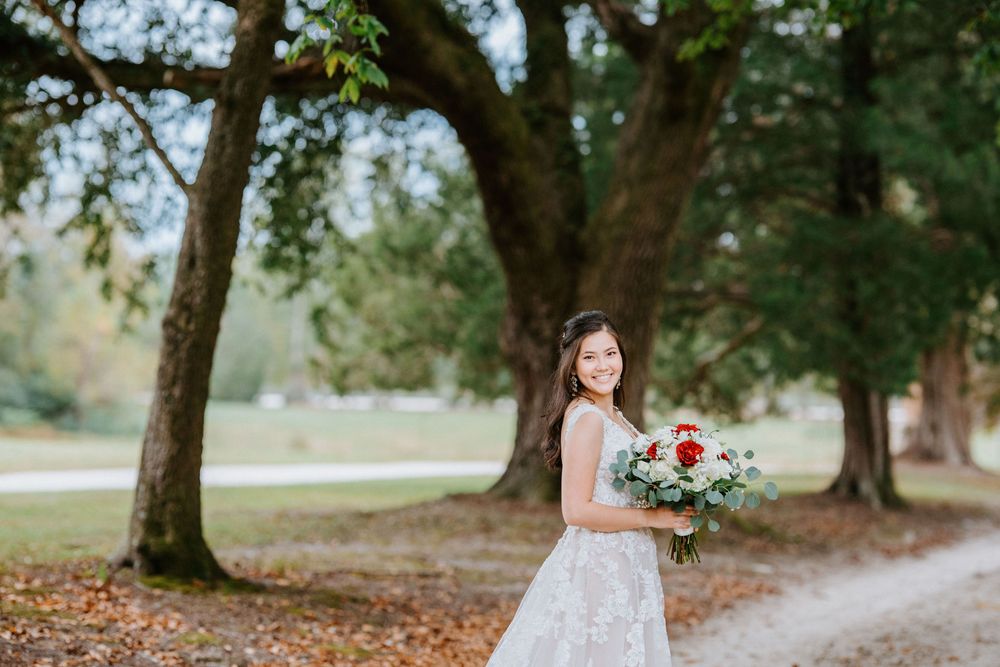 A bride in a white lace gown holds a vibrant red and white bouquet while standing under tall trees in a park setting.
