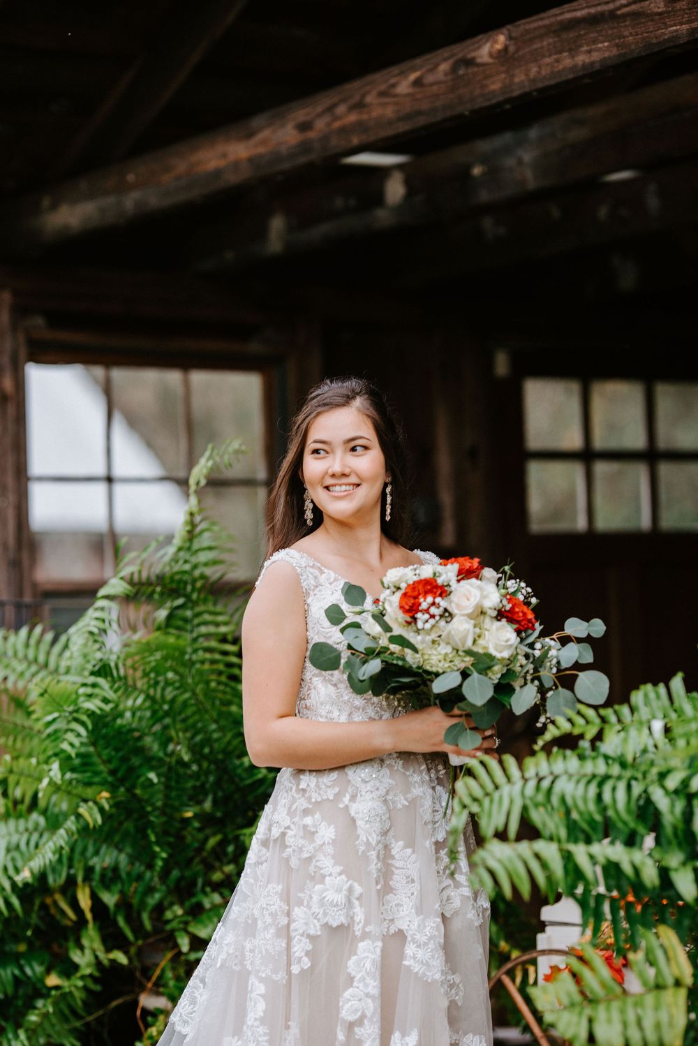 A bride wearing a lace wedding gown holds a red and white bouquet while standing near ferns and rustic wooden beams.