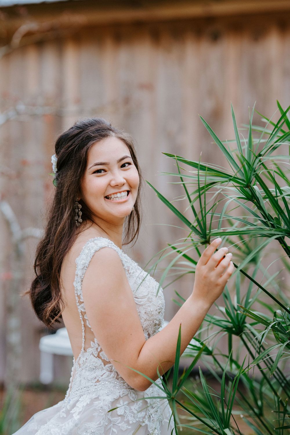A stunning lace wedding gown is displayed against a rustic barn backdrop with tropical foliage.