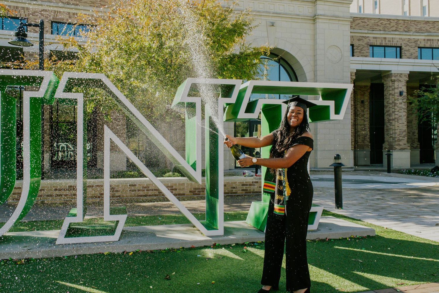College graduate poses near green university letters and architectural elements. UNT Graduate Portraits by Eric Aaberg Photography in DFW.