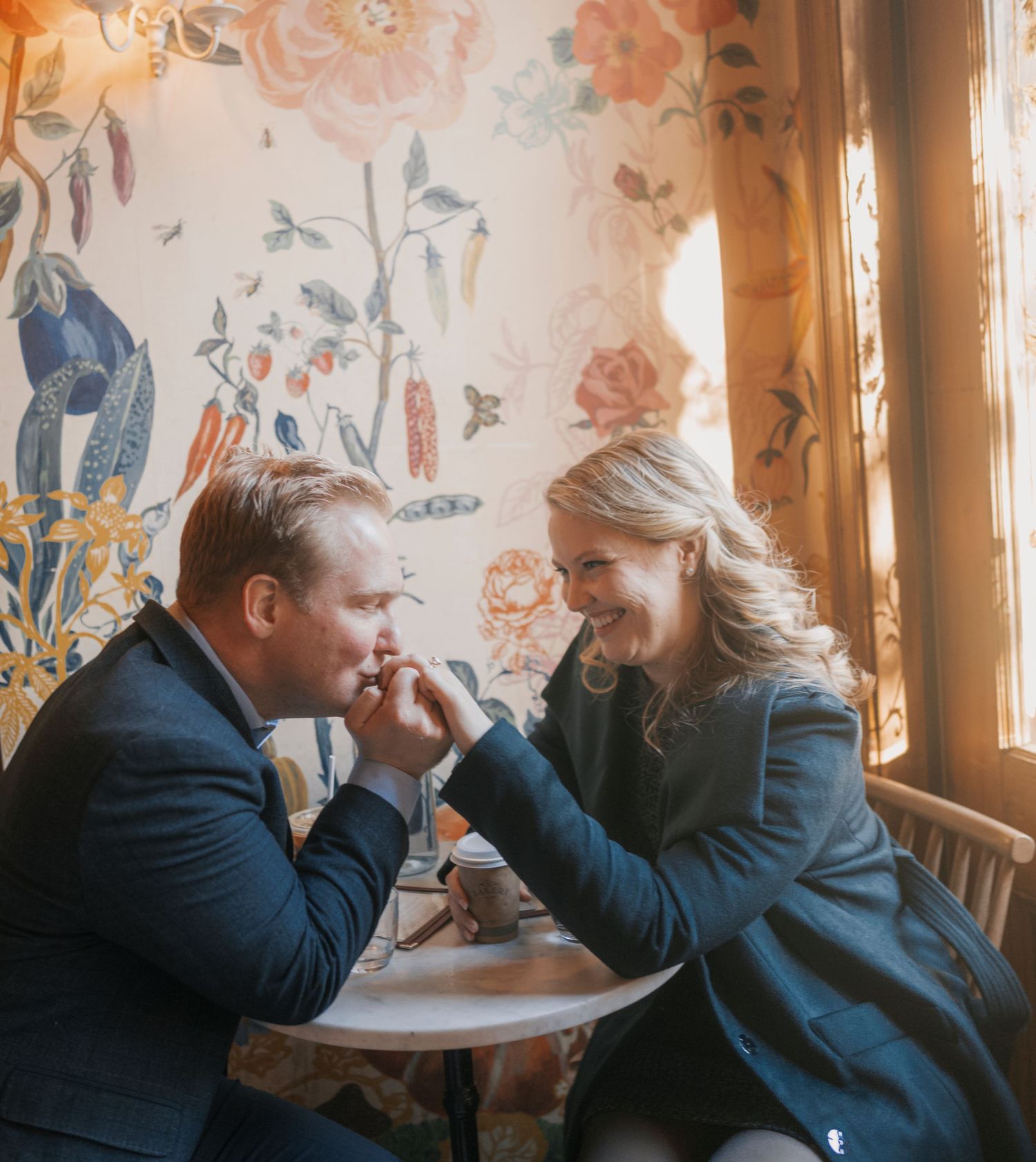 A couple shares an intimate moment at a cafe table with floral wallpaper and warm lighting in the background.